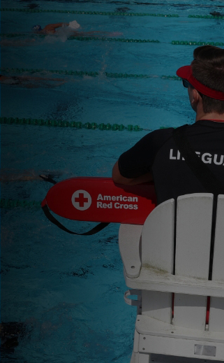 Image of a Lifeguard holding a Red Cross lifeguard rescue tube, sitting, and watching a pool full of people swimming laps.